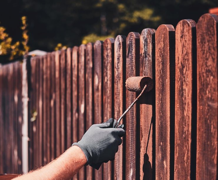 Person applying stain to a wooden fence with a roller, showcasing deck and fence maintenance in Florida's humid climate.